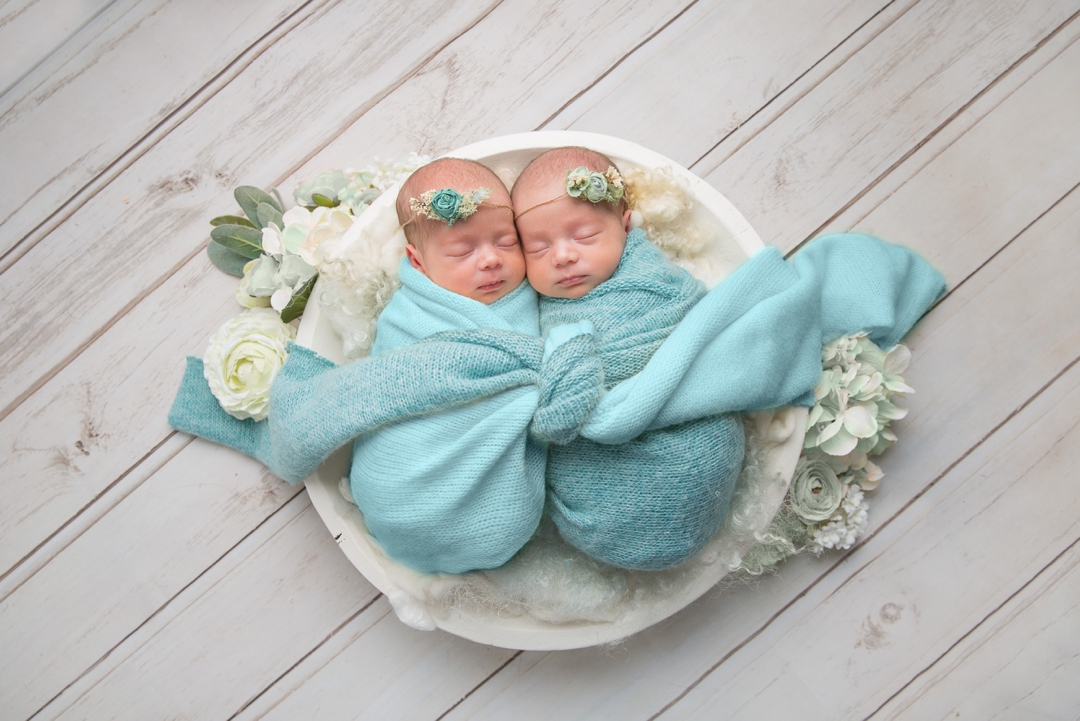 Newborn photography by hayley morris - image shows newborn baby twins wrapped in posing bowl together in coordinating colours on a wooden flower with flower head pieces and flowers surrounding them 